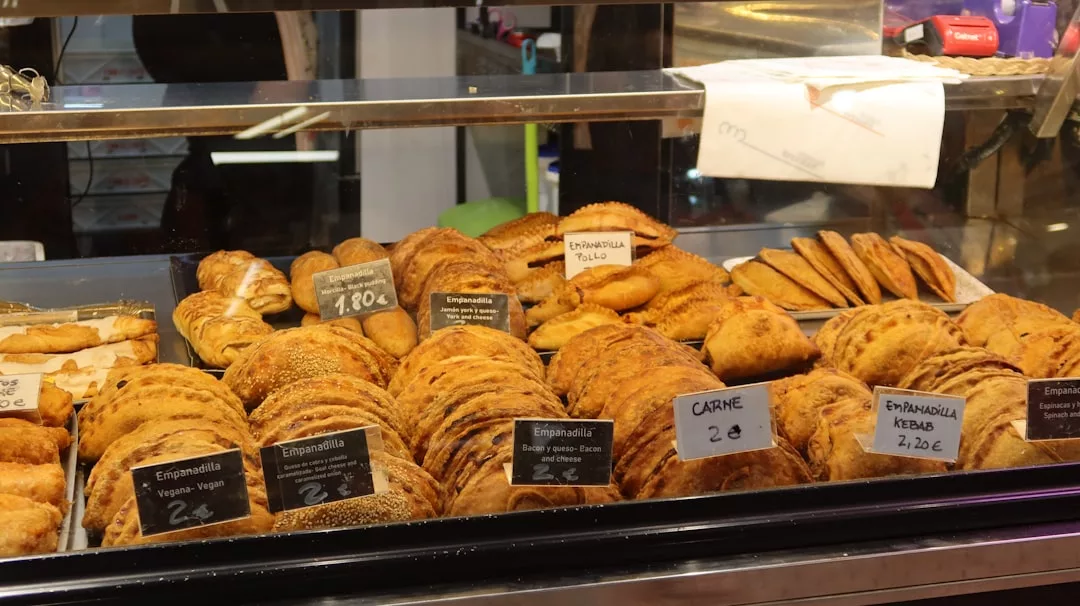Eine Vielzahl von Empanadas und Backwaren, darunter auch mallorquinisches Gebäck, werden in einer Bäckerei auf Tabletts präsentiert, vor denen handgeschriebene Preisschilder angebracht sind.