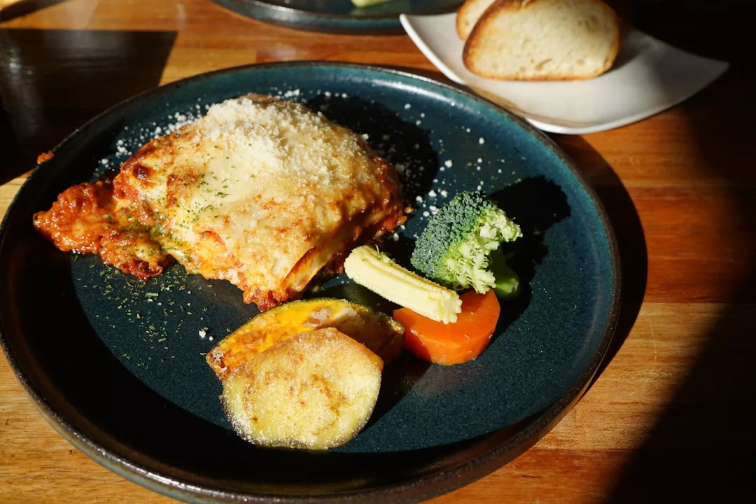 Ein Teller mit einer Portion Lasagne, überbacken mit geriebenem Käse, begleitet von Brokkoli, Mais, Karotten und Kartoffelscheiben - ein Geschmack von Mallorca -, serviert mit Brot auf einem separaten Teller im Hintergrund.
