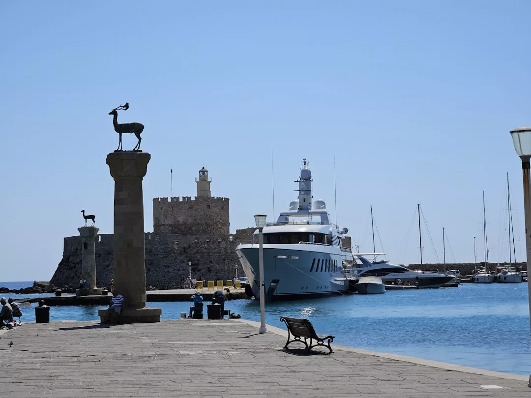 Ein steinerner Pier auf Mallorca mit einer Hirschstatue, einer Bank und sitzenden Menschen überblickt Yachten, die in einem Hafen in der Nähe einer alten Steinfestung angedockt sind.