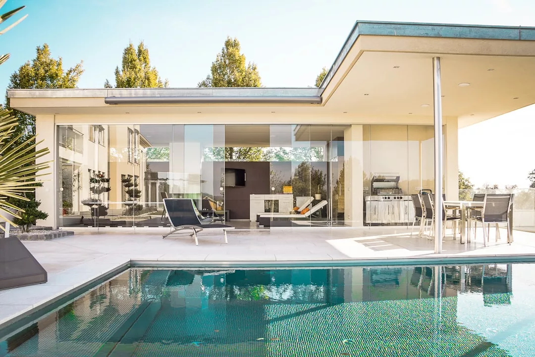 Modernes Haus auf Mallorca mit großen Glasfenstern, einem Innenhof mit Liegestühlen, einem Grill und einem Esstisch im Freien neben einem Swimmingpool.