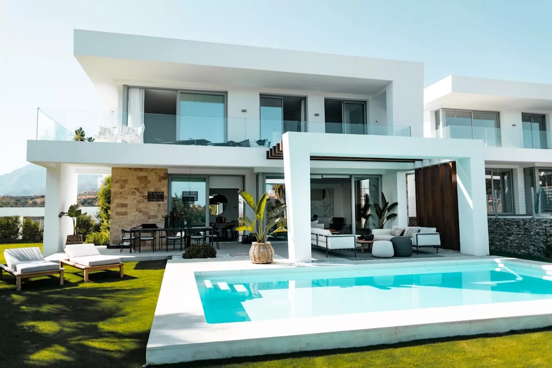 Modernes zweistöckiges weißes Haus auf Mallorca mit großen Fenstern, einem Sitzbereich im Freien, Sonnenliegen und einem Pool im Vordergrund an einem sonnigen Tag.