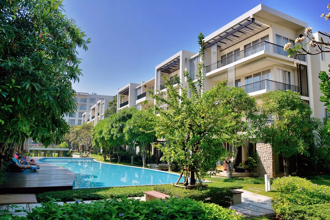 Die modernen Apartmentgebäude mit Balkonen blicken auf einen angelegten Garten und einen Swimmingpool im Freien, umgeben von Bäumen und Grün unter Mallorcas klarem blauen Himmel.