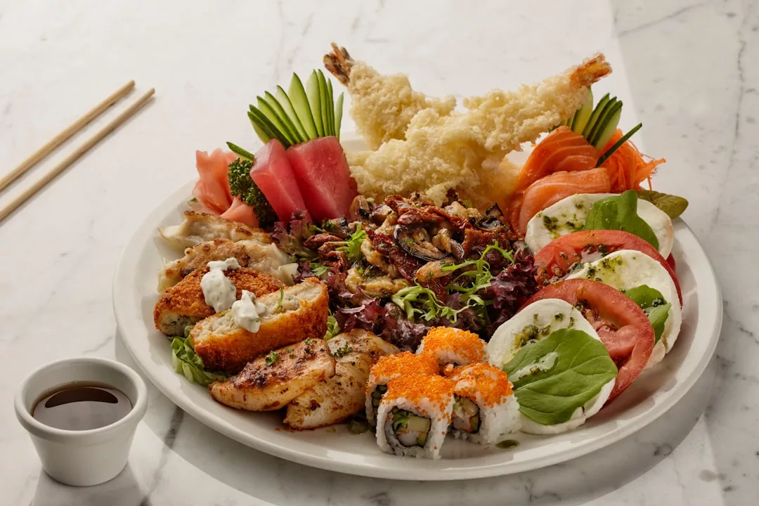 Eine große, von Mallorca inspirierte Platte mit verschiedenen Sushi-Rollen, Tempura-Garnelen, Sashimi, Hühnchen, Gyoza, Salat und Tomatenscheiben mit Mozzarella und Basilikum, serviert mit einer kleinen Schale Sojasauce.