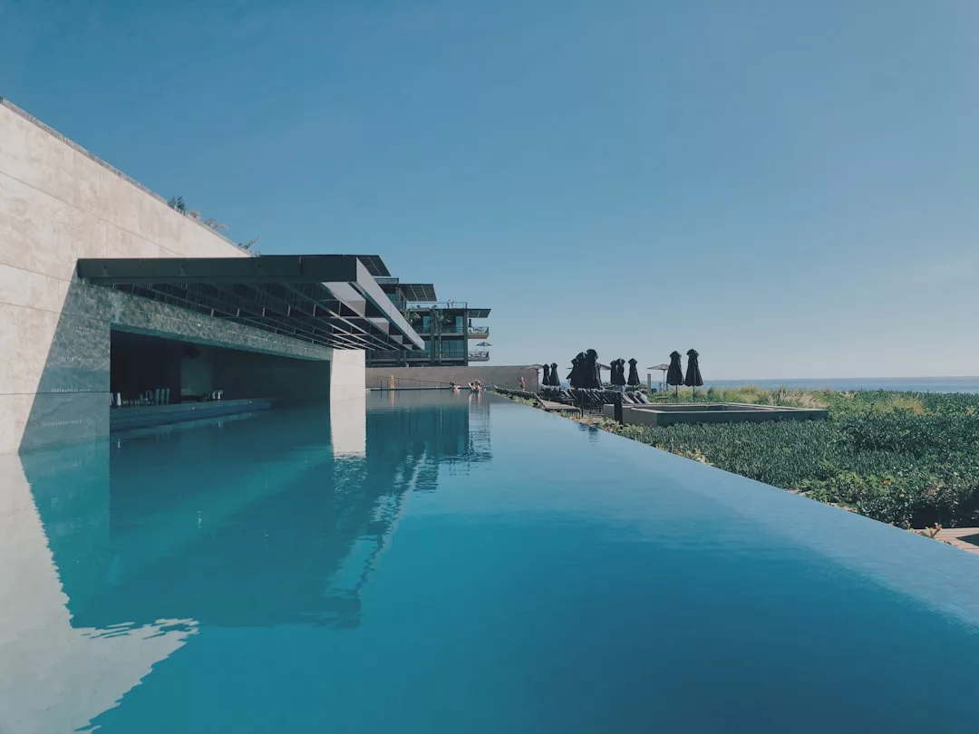 Infinity-Pool mit klarem, blauem Wasser neben einem modernen Gebäude auf Mallorca, mit Menschen auf einer Terrasse im Hintergrund unter klarem Himmel.