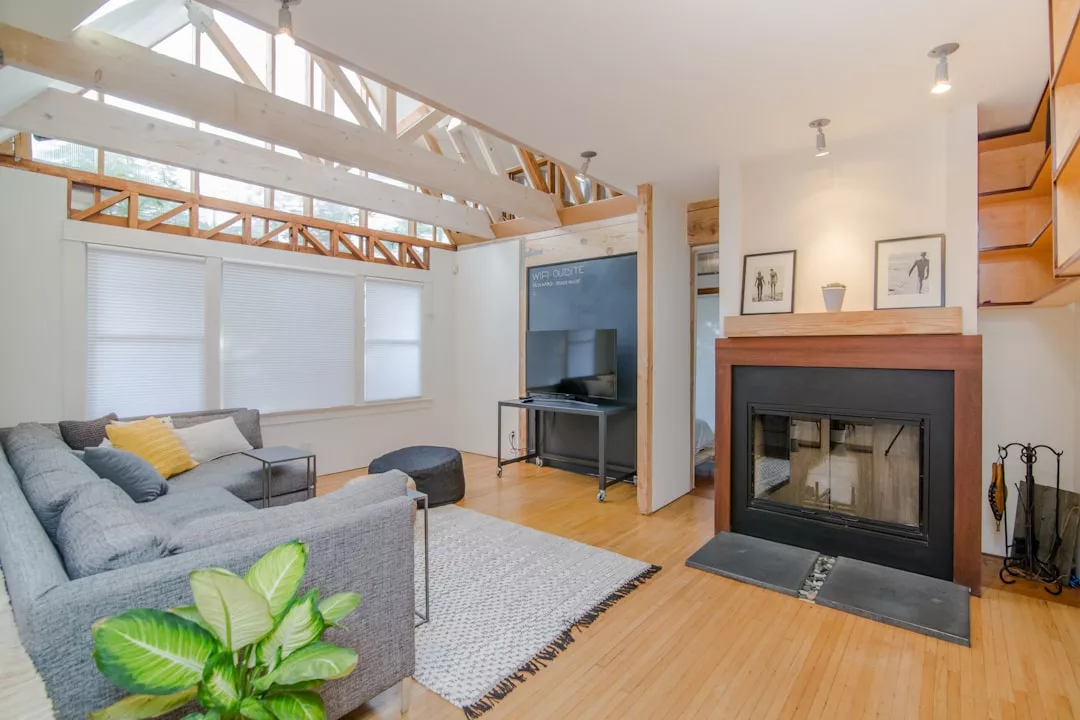 Ein modernes mallorquinisches Wohnzimmer mit einem grauen Sektionssofa, einem Kamin, einem Fernseher, Holzböden, Sichtbalken und großen Fenstern mit Jalousien.