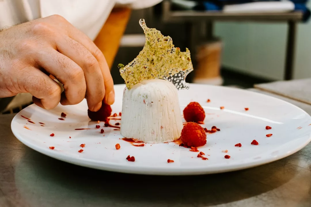 Eine Person arrangiert Himbeeren und zerkleinerte rote Garnierung um ein geformtes weißes Mallorca-Dessert, das mit einem dekorativen Crisp auf einem weißen Teller gekrönt ist.