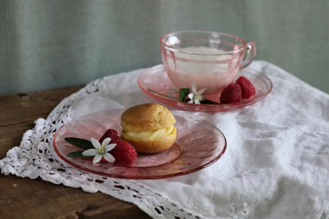 Auf einem rosafarbenen Glasteller, der an den süßen Charme Mallorcas erinnert, ruht ein Windbeutel mit Himbeeren und kleinen weißen Blüten neben einer rosafarbenen Glasteetasse auf einem spitzenbesetzten weißen Tuch.