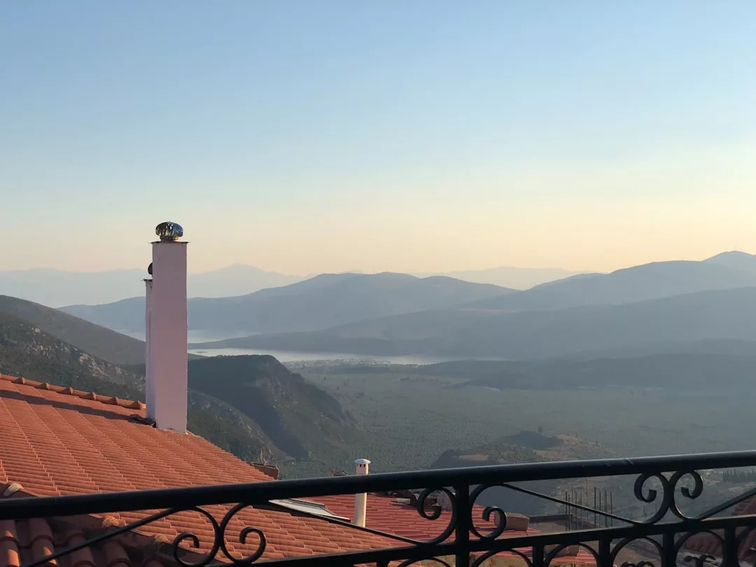 Blick auf die roten Ziegeldächer Mallorcas, einen weißen Schornstein und ein Metallgeländer im Vordergrund, mit sanften Hügeln und einem entfernten See unter einem klaren Himmel im Hintergrund.
