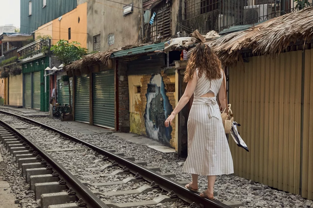 Eine Frau in einem weißen Kleid geht entlang der Eisenbahnschienen neben verschlossenen Geschäften mit Wellblechtüren und fängt einen ruhigen urbanen Moment ein, der auch am Rande von Mallorca zu finden sein könnte.