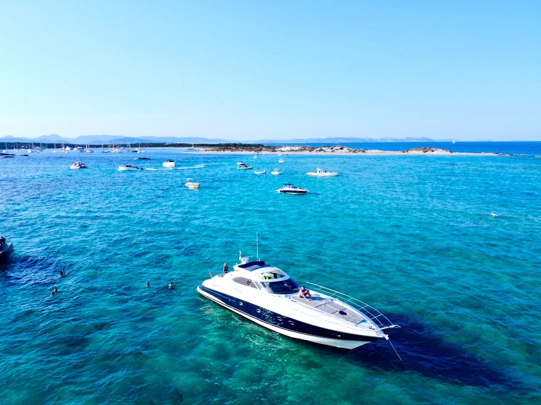 Eine weiß-blaue Motoryacht schwimmt auf dem klaren türkisfarbenen Wasser in der Nähe von Mallorca, umgeben von mehreren kleineren Booten, mit einer felsigen Küstenlinie und fernen Hügeln unter einem klaren blauen Himmel.