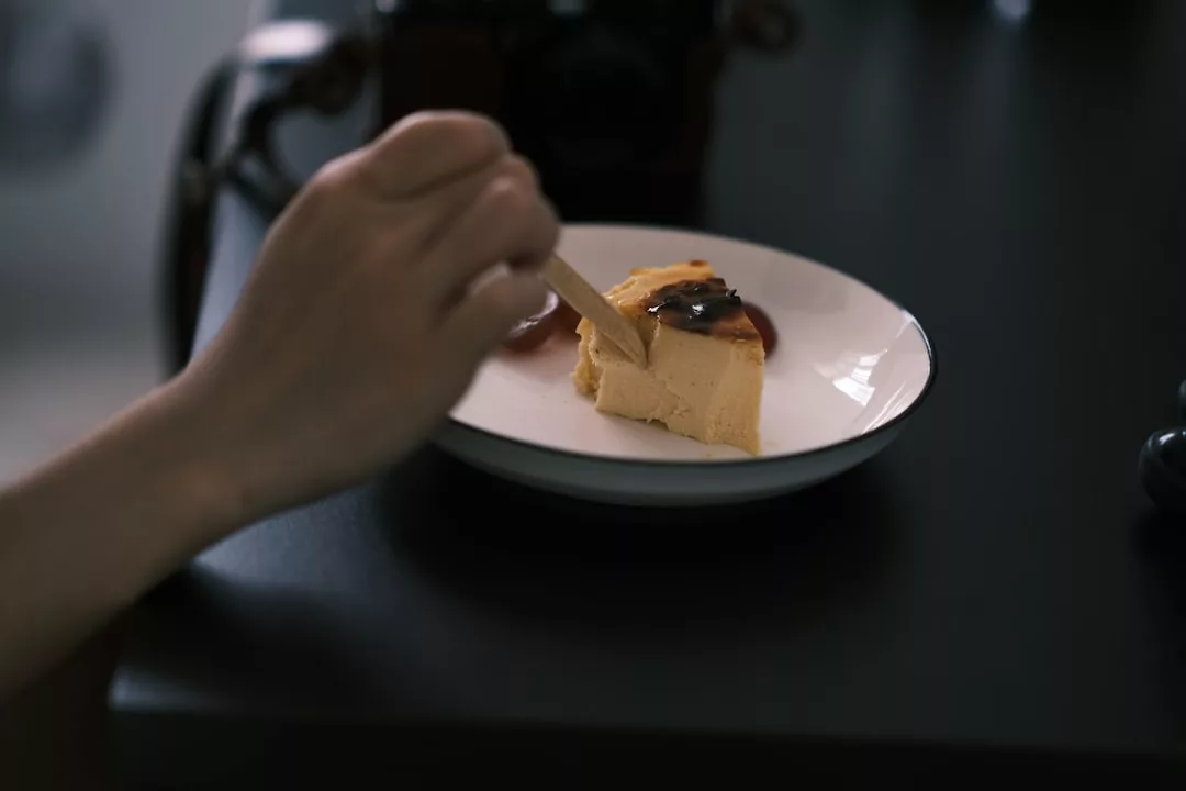 Eine Hand hält eine Gabel und ist dabei, ein Stück Kuchen mit dunklem Sirup auf einem weißen Teller zu essen, der auf einem dunklen Tisch auf Mallorca steht, mit einer unscharfen Kamera im Hintergrund.