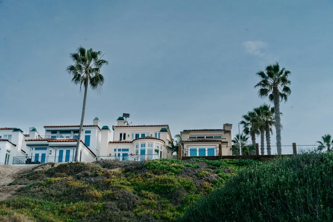 Ein großes Küstenhaus mit weißen Wänden und blauen Verzierungen liegt auf einem grasbewachsenen Hang auf Mallorca, umgeben von Palmen unter einem klaren blauen Himmel.