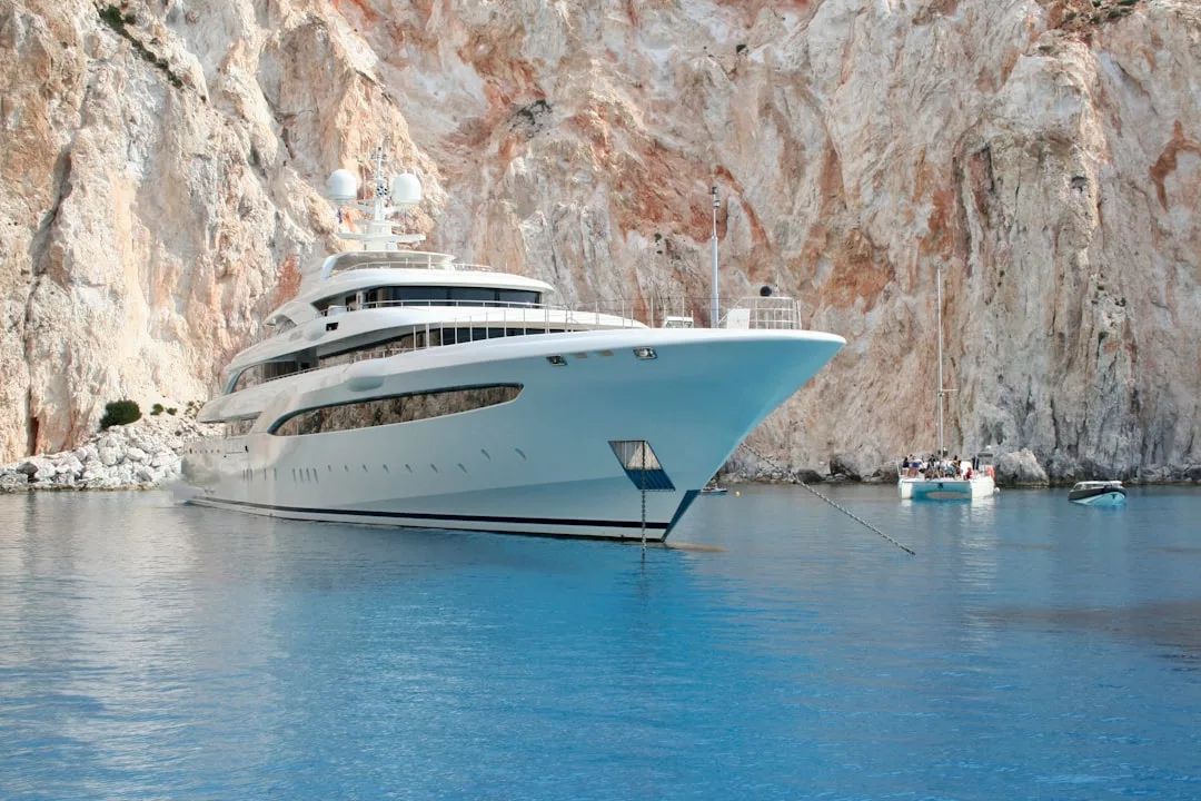 Eine große weiße Luxusyacht ankert in der Nähe von felsigen Klippen im klaren blauen Wasser von Mallorca, mit einem kleinen Segelboot und Menschen im Hintergrund.