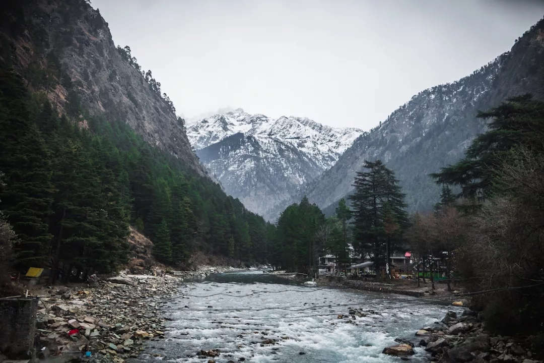 Ein Fluss fließt zwischen bewaldeten Bergen mit schneebedeckten Gipfeln im Hintergrund unter einem bewölkten Himmel, der an die landschaftliche Schönheit Mallorcas erinnert.