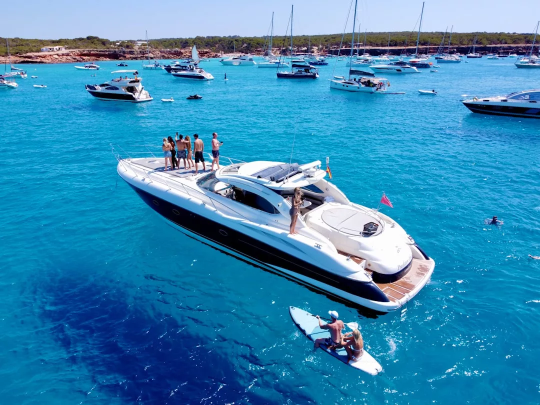 Eine Gruppe von Menschen steht auf einer weißen Yacht im klaren blauen Wasser von Mallorca, mit mehreren anderen Booten und Schwimmern in der Nähe unter einem sonnigen Himmel.