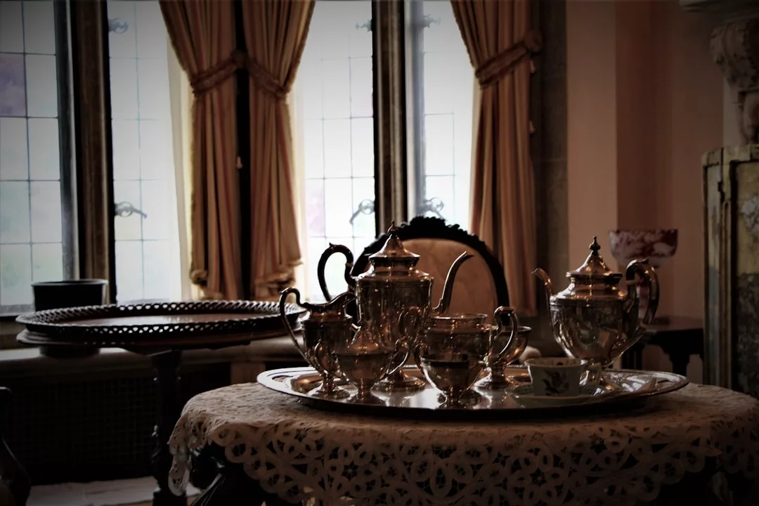 Ein silbernes Teeservice liegt auf einem verzierten Tablett auf einem mit Spitze bedeckten Tisch vor großen, mit Vorhängen versehenen Fenstern und bringt einen Hauch von mallorquinischer Eleganz in den traditionell eingerichteten Raum.