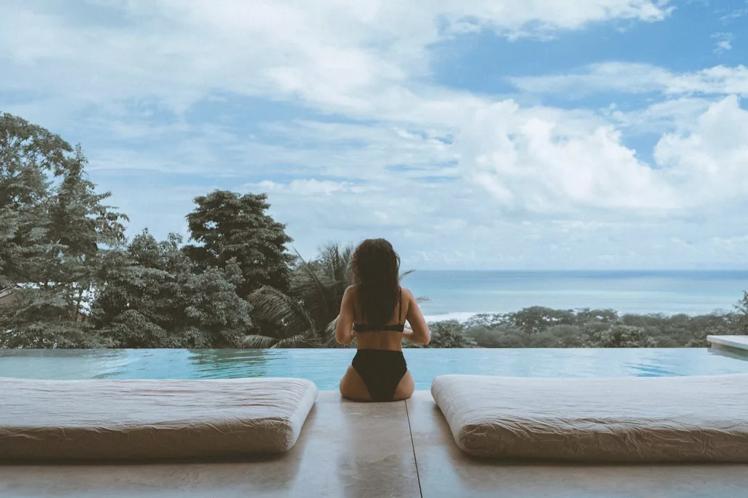 Eine Frau in einem schwarzen Badeanzug sitzt am Rande eines Infinity-Pools auf Mallorca und blickt auf üppige Bäume und das Meer unter einem teilweise bewölkten Himmel.