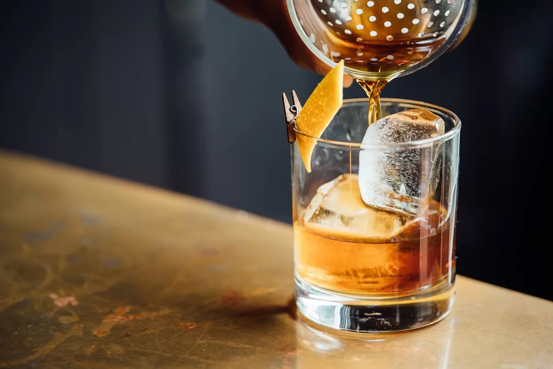 In einer Bar auf Mallorca wird ein Cocktail durch ein Sieb in ein Glas mit Eis und einer Zitronenzeste gegossen.