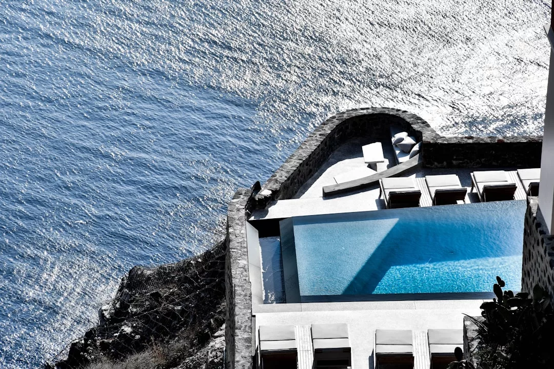 Ein rechteckiger Swimmingpool und Sonnenliegen überblicken ein glitzerndes mallorquinisches Gewässer, mit Steinmauern und einem schattigen Sitzbereich in der Nähe.