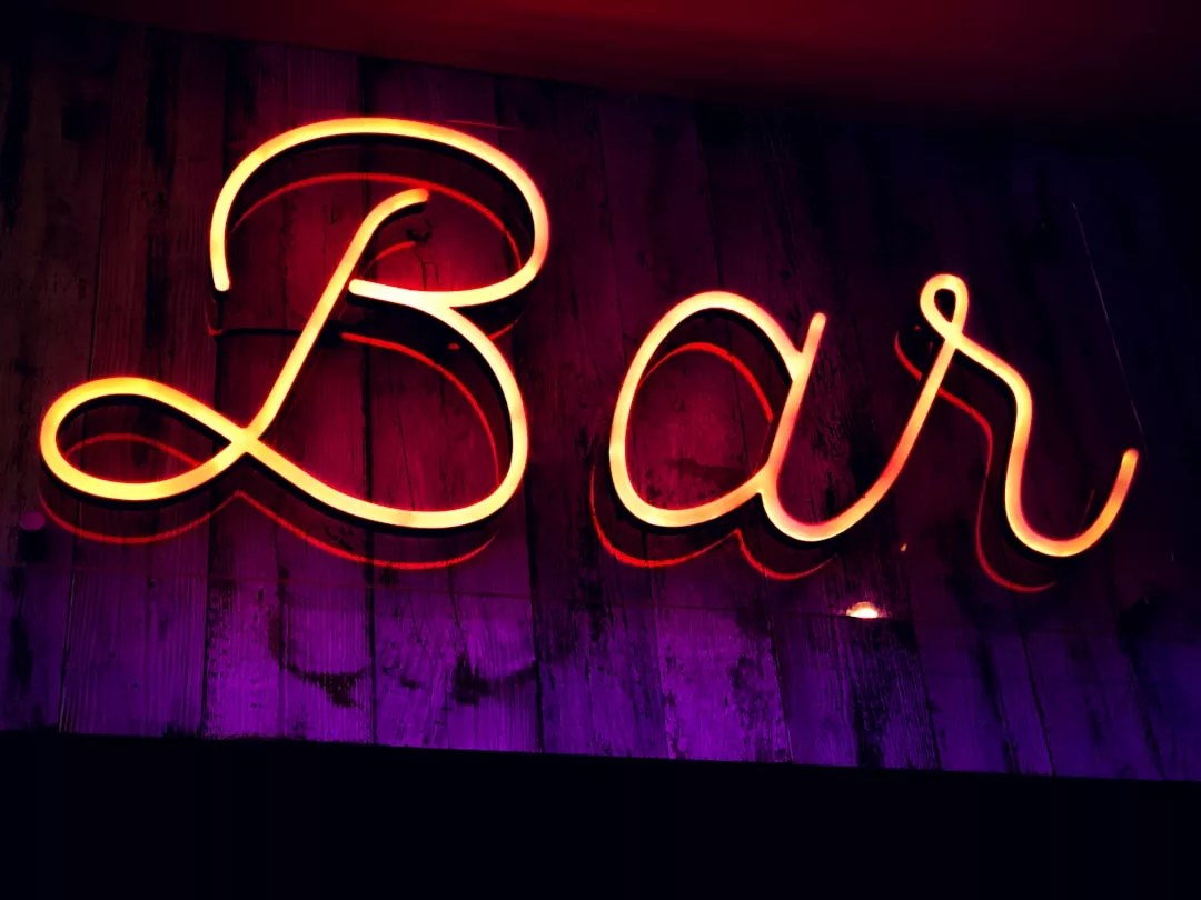 Ein Neonschild mit dem Schriftzug "Bar" in kursiven Buchstaben, beleuchtet in Gelb und Orange vor einer dunklen Holzwand mit violetter Beleuchtung, schafft eine lebendige Atmosphäre im Nachtleben von Mallorca.