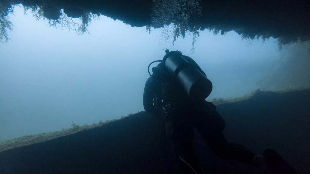 Ein Taucher schwimmt in der Nähe des Randes einer Unterwasserhöhle auf Mallorca, während das Sonnenlicht durch das Wasser darüber fällt.