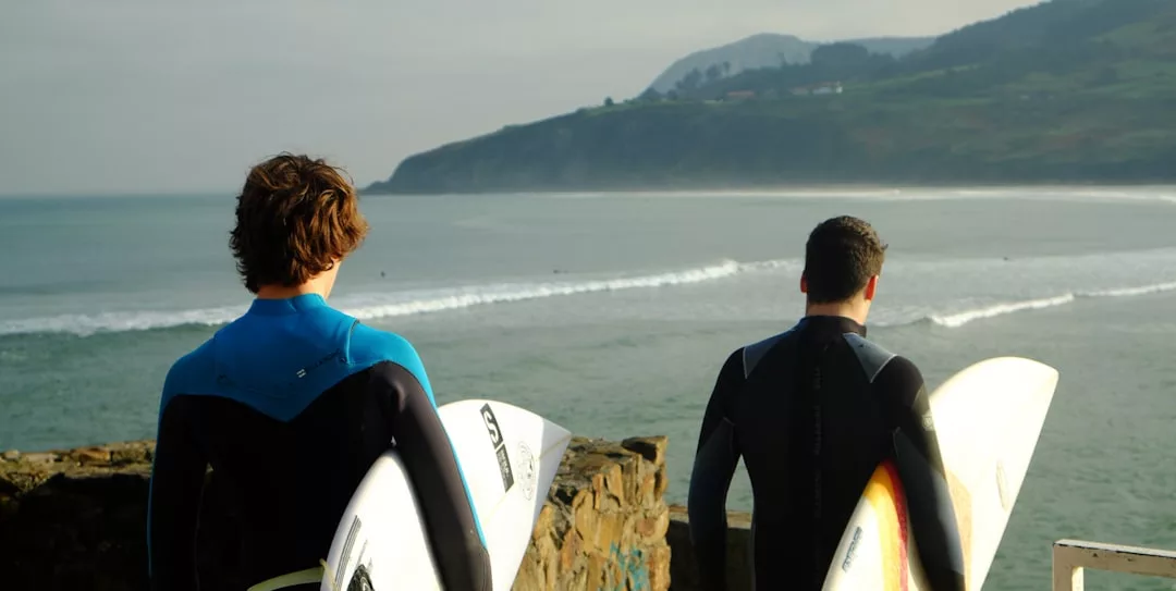 Zwei Surfer in Neoprenanzügen und mit Surfbrettern in der Hand stehen auf einem Steinsockel und blicken auf das Meer mit den Wellen und den malerischen Hügeln Mallorcas im Hintergrund.