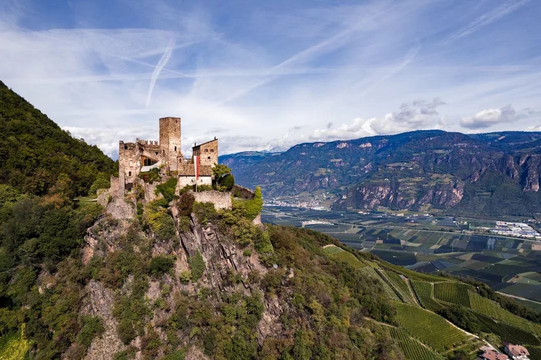 Ein steinernes Schloss steht auf einem felsigen Hügel auf Mallorca, umgeben von grünen Weinbergen und Bergen unter einem blauen Himmel mit Wolkenschlieren.