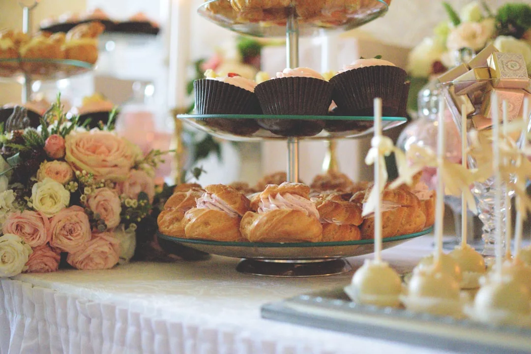 Ein Desserttisch mit Gebäck, Cupcakes und Cake Pops auf gestapelten Tabletts, inspiriert von Mallorca, umgeben von Blumenarrangements und einer weißen Tischdecke.
