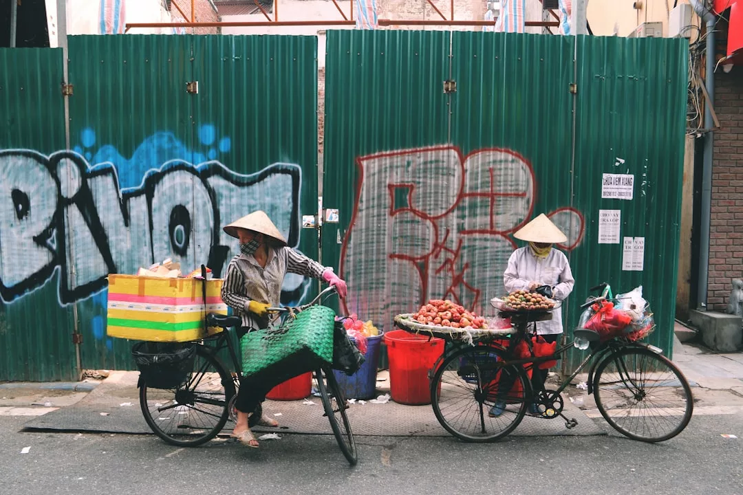 Zwei Straßenhändler mit kegelförmigen Hüten verkaufen Obst von Fahrrädern aus vor einem grünen Metallzaun mit Graffiti und bringen so einen Hauch von Mallorcas pulsierender Marktkultur auf einen städtischen Bürgersteig.