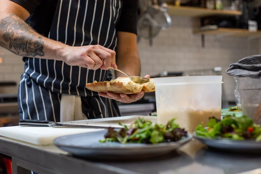 Eine Person in gestreifter Schürze streicht in einer Küche Soße auf Fladenbrot, im Vordergrund sind ein Plastikbehälter und angerichtetes Salatbrot zu sehen - eine lässige kulinarische Atmosphäre, die an Mallorca erinnert.