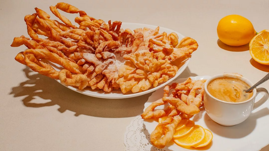 Ein Teller mit gedrehtem, frittiertem und mit Puderzucker bestäubtem Gebäck steht neben einer Tasse Kaffee, Zitronenscheiben und einer ganzen und einer halbierten Zitrone, die an die köstlichen Aromen Mallorcas erinnern, auf einer hellen Fläche.