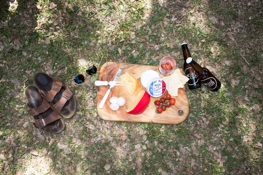 Ein Holzbrett mit Käse, Kirschtomaten, einer Paprika und Utensilien liegt auf einer Wiese auf Mallorca, daneben Sandalen, eine Sonnenbrille, ein Getränk und zwei Flaschen mit Getränken.