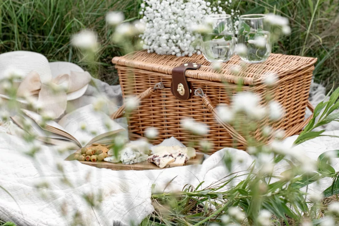 Ein Picknickkorb aus Weidengeflecht, zwei Gläser, ein Teller mit Essen und ein Strauß weißer Blumen liegen auf einer Decke, umgeben von Gras und Grün, und erinnern an den entspannten Charme Mallorcas.