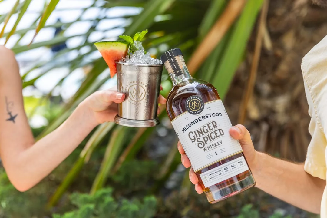 Eine Person hält eine Flasche Misunderstood Ginger Spiced Whiskey in der Hand, während eine andere einen mit Eis, Minze und einer Wassermelonenscheibe gefüllten Metallbecher in die Höhe hält - perfekt, um die lebhaften Aromen Mallorcas zu genießen.