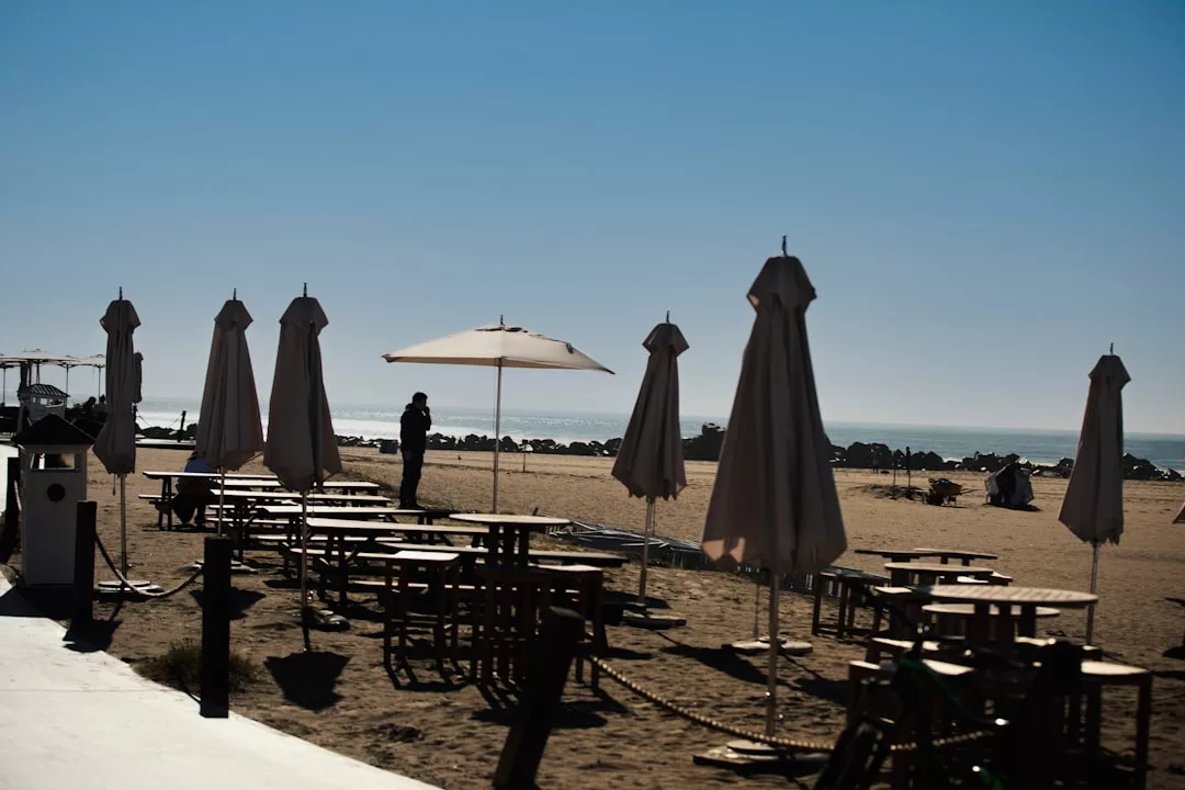 Mehrere geschlossene Terrassenschirme und leere Picknicktische stehen an einem Sandstrand auf Mallorca unter strahlend blauem Himmel. Im Hintergrund steht eine Person in der Nähe des Ufers.