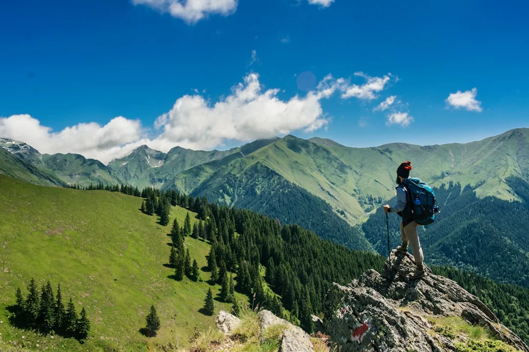 Ein Wanderer mit Rucksack steht auf einem Felsvorsprung auf Mallorca und blickt auf grüne Berge und einen Wald unter einem blauen, wolkenverhangenen Himmel.
