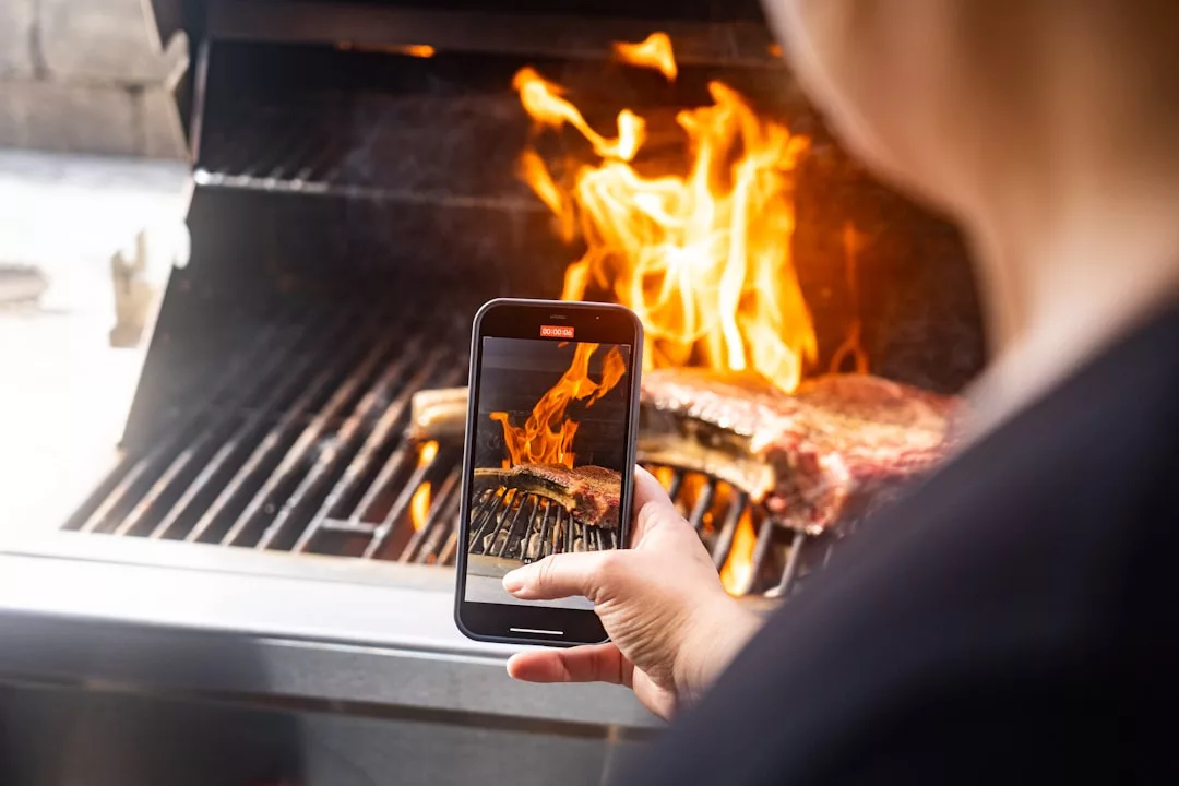 Eine Person fotografiert mit ihrem Smartphone einen brennenden Grill, auf dem Fleisch gegart wird, und genießt die lebhafte Atmosphäre auf Mallorca.
