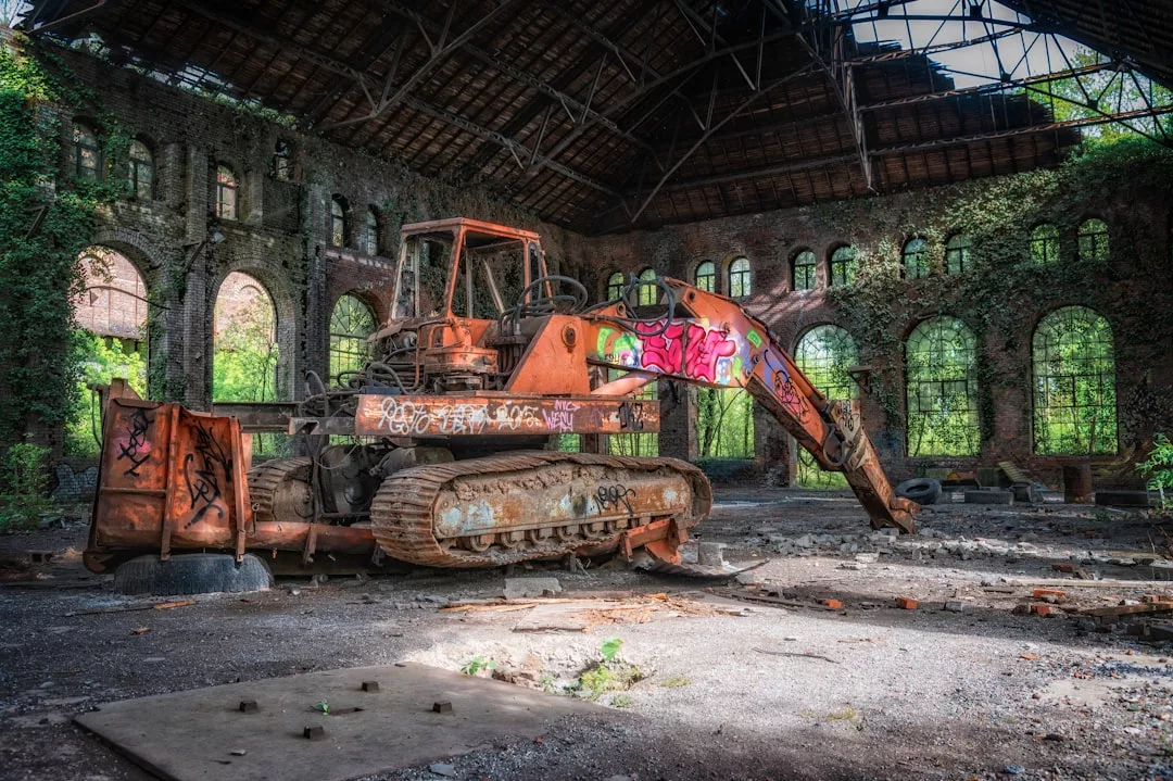 Ein alter, verrosteter und mit Graffiti beschmierter Bagger steht in einem großen, verlassenen Gebäude mit zerbrochenen Fenstern und überwucherter Vegetation, das auf die verborgene, vergessene industrielle Vergangenheit Mallorcas hinweist.