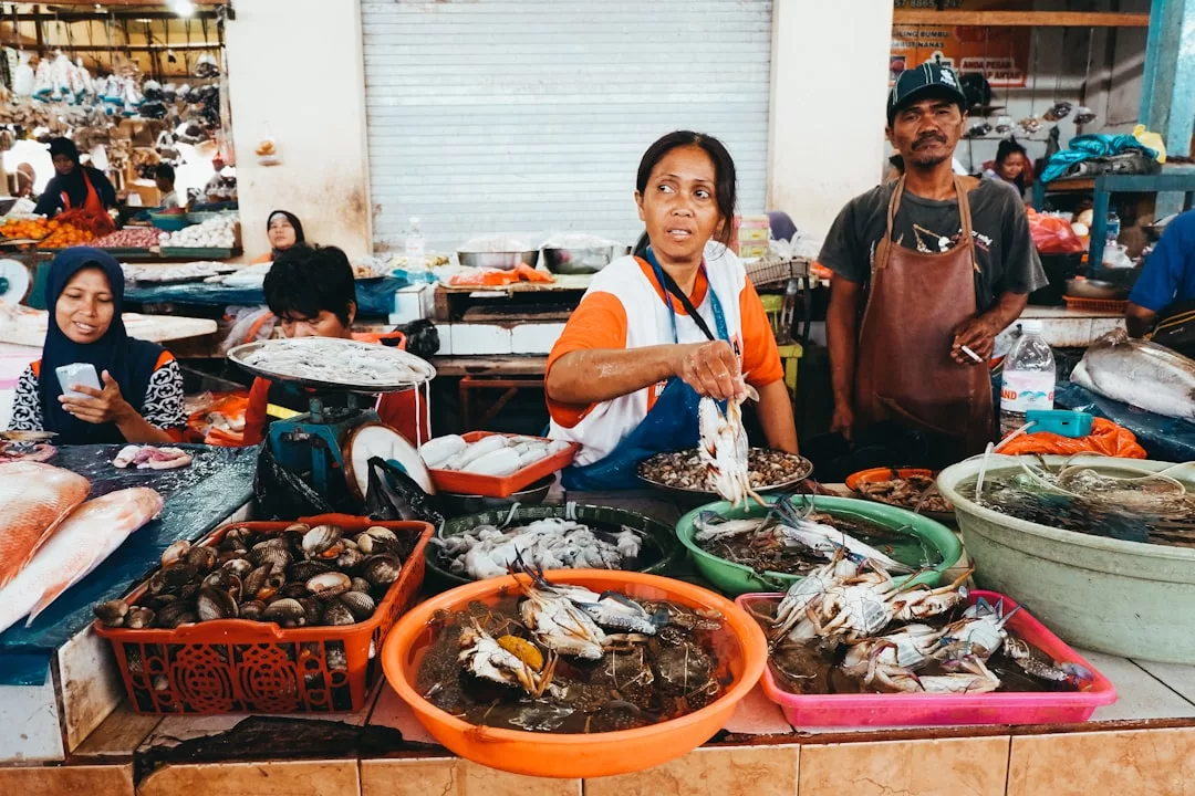 Verkäufer auf einem belebten Markt auf Mallorca verkaufen frische Meeresfrüchte, darunter Krabben und Fisch, die in Schalen angeboten werden. Man sieht Menschen, die hinter der Theke arbeiten und sitzen.