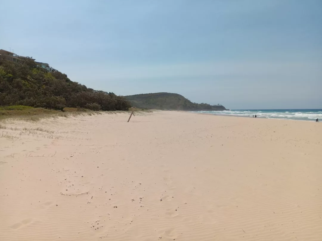 Ein breiter, leerer Sandstrand auf Mallorca erstreckt sich in Richtung Meer mit sanften Wellen und einem grasbewachsenen Hügel im Hintergrund unter einem klaren Himmel.
