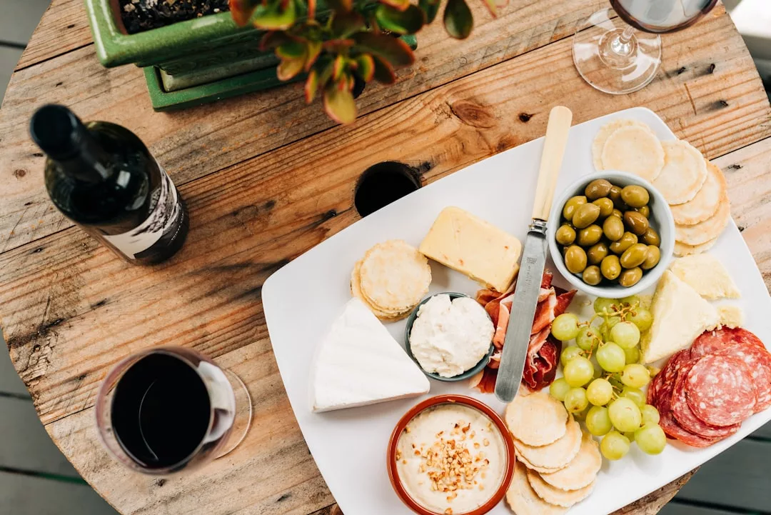 Ein Charcuterie-Brett mit verschiedenen Käsesorten, Crackern, grünen Oliven, Weintrauben, Wurstwaren und Dips ist wunderschön auf einem Holztisch auf Mallorca arrangiert, neben zwei Gläsern und einer Flasche Rotwein.