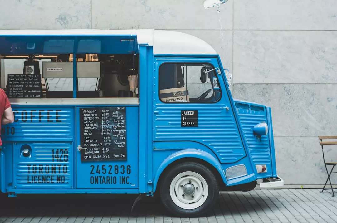 Ein blauer Kaffeewagen mit einer Speisekarte an der Seite ist auf einem gepflasterten Bürgersteig in Mallorca neben einer grauen Wand geparkt; ein Teil einer Person und ein Stuhl sind zu sehen.