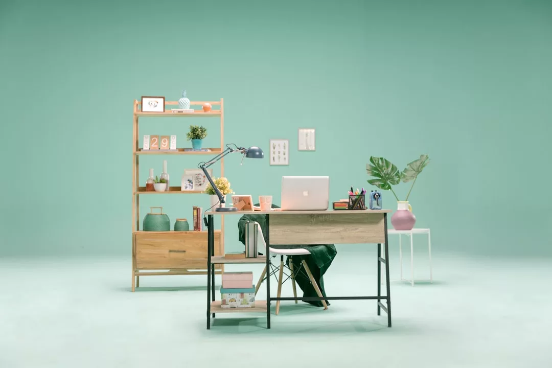 Ein ordentlich aufgeräumter Schreibtisch mit Laptop, Lampe und Schreibwaren steht vor einem Holzregal mit Büchern und Dekoration, das vor einem mintgrünen Hintergrund den entspannten Charme Mallorcas versprüht.