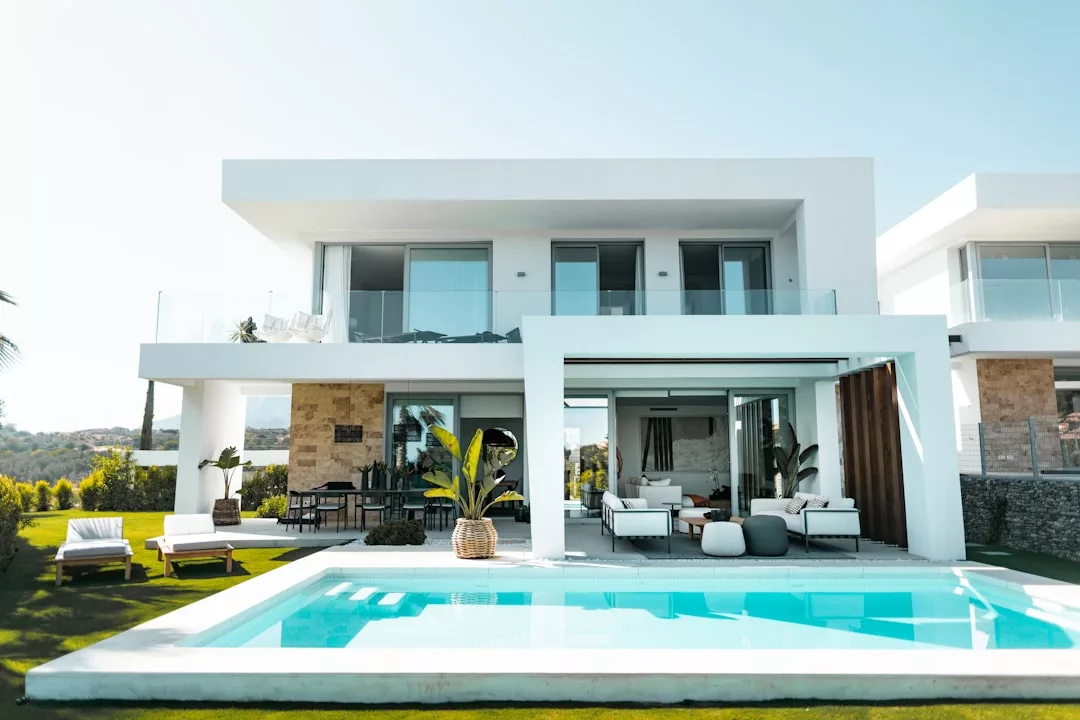 Modernes zweistöckiges Haus auf Mallorca mit großen Fenstern, Außenwohnbereich, Swimmingpool und Sonnenliegen auf einer gepflegten Rasenfläche.