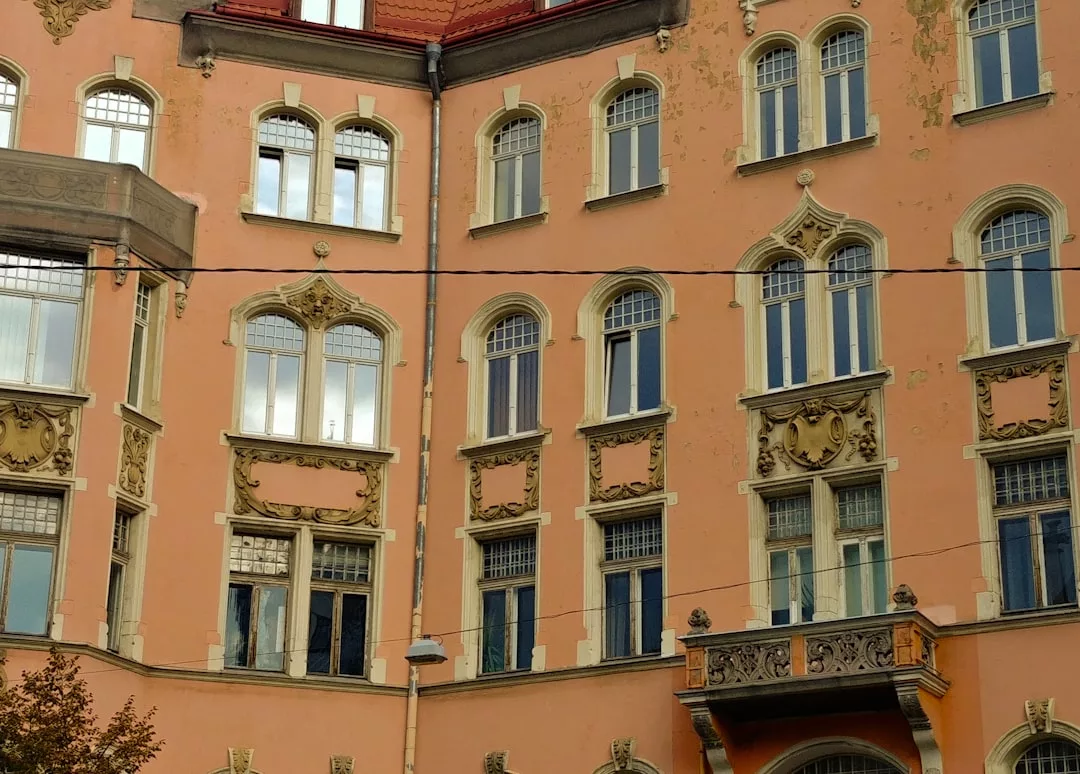 Verschnörkelte pfirsichfarbene Gebäudefassade auf Mallorca mit bogenförmigen und rechteckigen Fenstern, Zierleisten und einem kleinen Balkon. Einige Außenfarbe scheint verwittert.