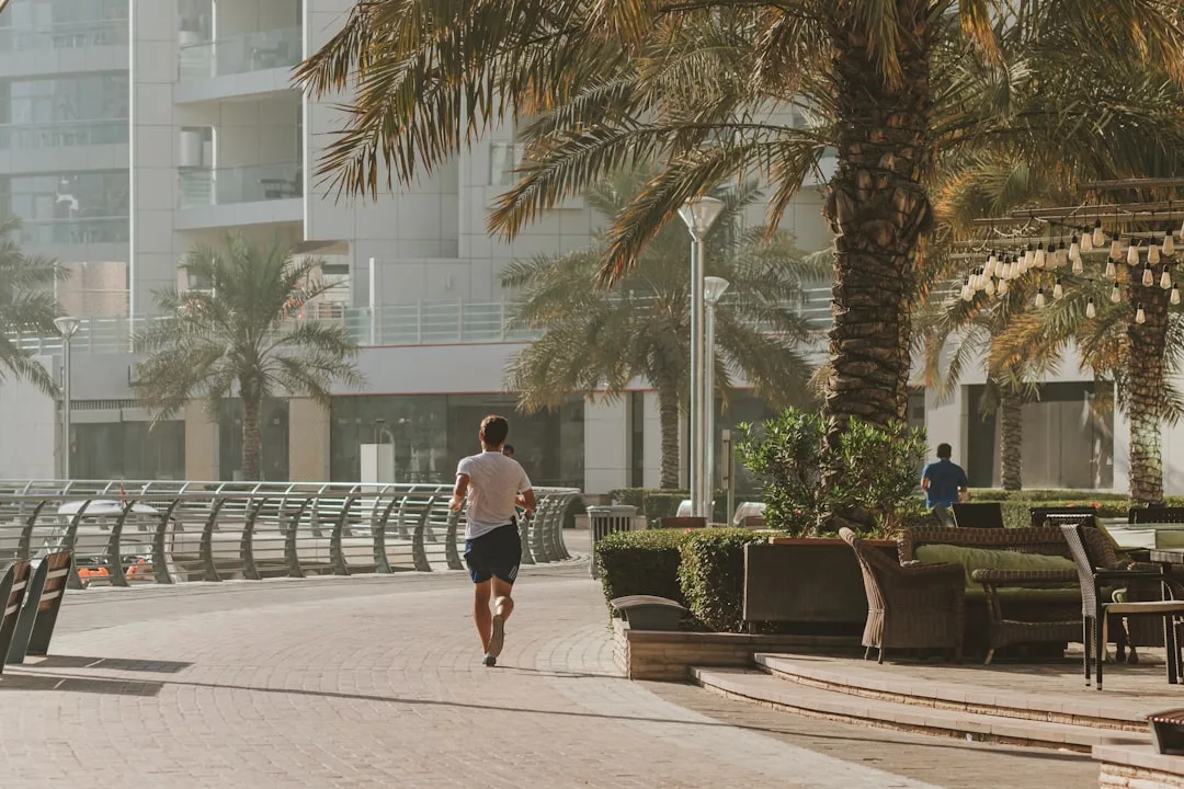 Eine Person joggt an einem sonnigen Tag auf Mallorca über einen gepflasterten Weg am Wasser, der von Palmen und Sitzgelegenheiten im Freien gesäumt ist.