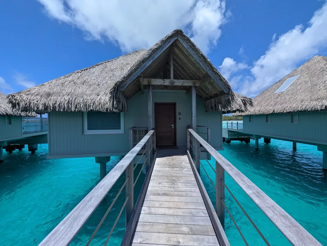 Ein Holzsteg führt zu einem strohgedeckten Überwasserbungalow, der über dem klaren türkisfarbenen Wasser unter einem teilweise wolkenverhangenen blauen Himmel erbaut wurde und den heiteren Charme eines versteckten Rückzugsortes auf Mallorca versprüht.