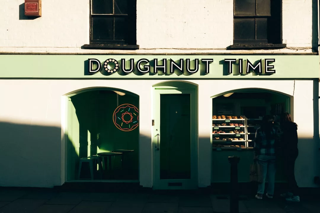 Ein Laden mit einem grünen Schild mit der Aufschrift "Doughnut Time" über gewölbten Fenstern und einer Tür, die an mallorquinische Cafés erinnert, in denen Krapfen ausgestellt sind und zwei Personen davor stehen.