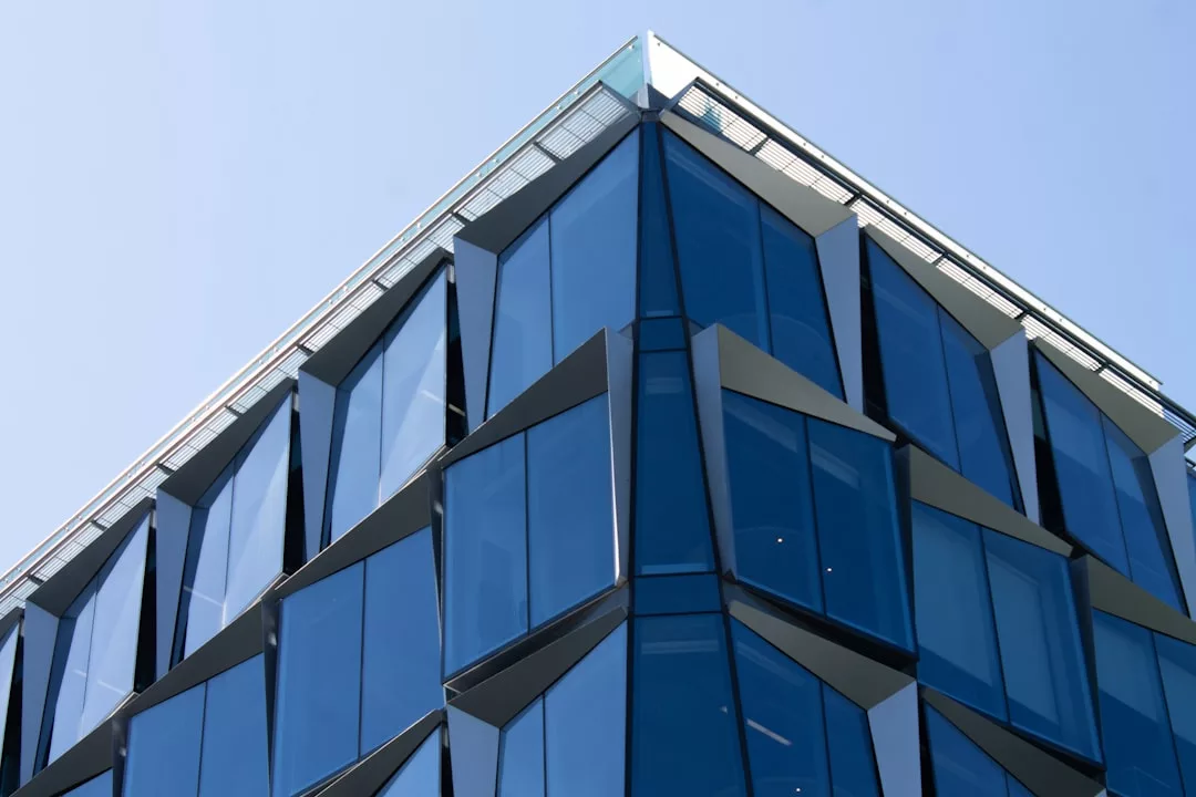 Ein modernes Gebäude auf Mallorca mit geometrischen, eckigen Glasfenstern, in denen sich der Himmel spiegelt, aus einem niedrigen Winkel vor einem klaren blauen Hintergrund betrachtet.