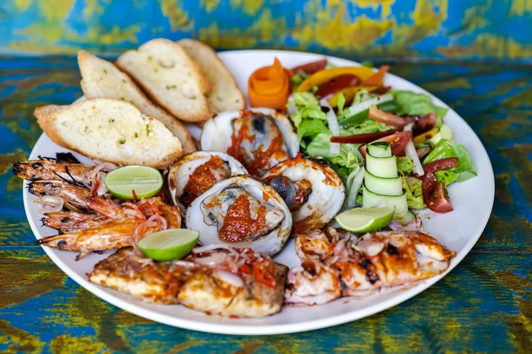 Ein Teller mit gegrillten Meeresfrüchten, aufgeschnittenem Brot, Limettenspalten, gemischtem Salat und Gurkenspiralen auf einer weißen Platte erinnert an die lebhaften Aromen Mallorcas vor einem farbenfrohen Hintergrund.
